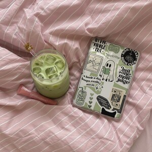 May include: A refreshing green drink with ice cubes in a clear glass, a tube of lip balm, and a phone case with stickers on a pink striped bedsheet. The phone case has text that says "ROMANTAS BOOK CLUB".