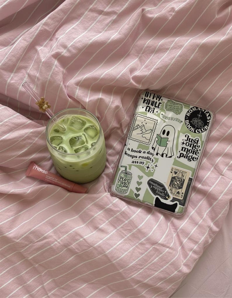 May include: A refreshing green drink with ice cubes in a clear glass, a tube of lip balm, and a phone case with stickers on a pink striped bedsheet. The phone case has text that says "ROMANTAS BOOK CLUB".