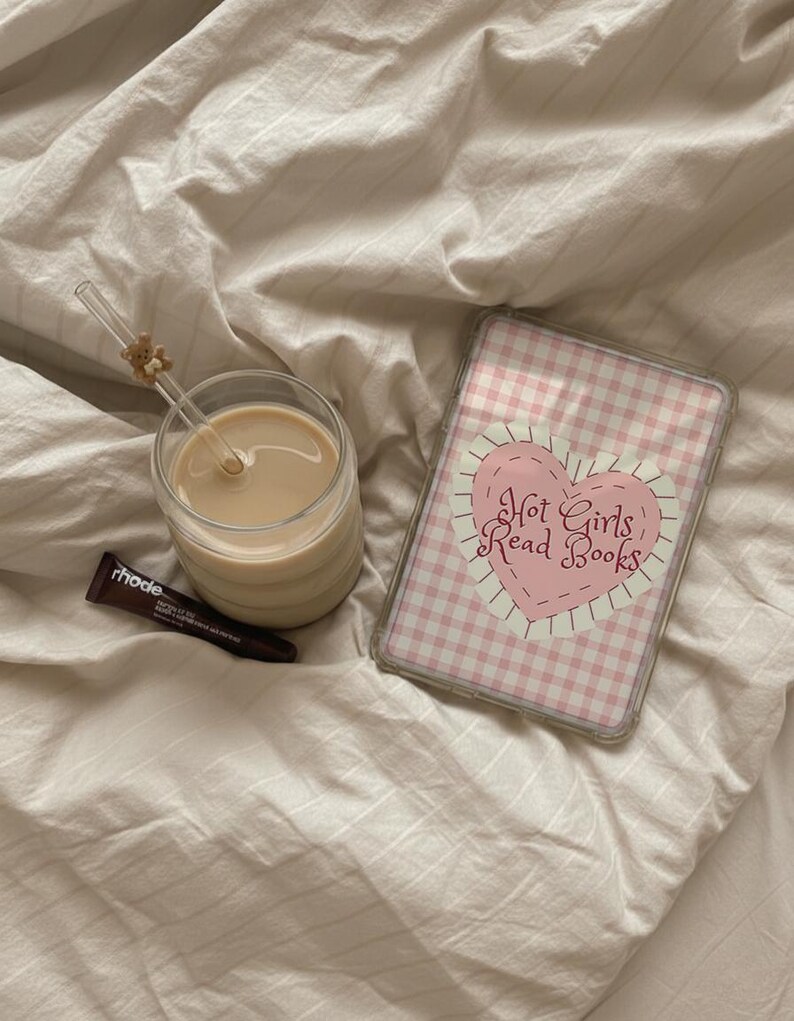 May include: A glass of coffee with a straw and a small teddy bear decoration, a tube of rhode lip treatment, and a phone case with a pink gingham pattern and the text "Hot Girls Read Books."