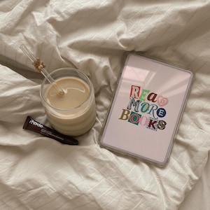 May include: A clear glass of coffee with a straw and a small teddy bear charm, a tablet with the words "READ MORE BOOKS" in colorful letters, and a tube of rhode lip treatment on a white bedsheet.