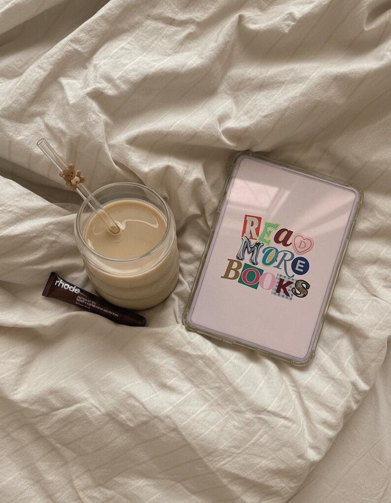 May include: A clear glass of coffee with a straw and a small teddy bear charm, a tablet with the words "READ MORE BOOKS" in colorful letters, and a tube of rhode lip treatment on a white bedsheet.
