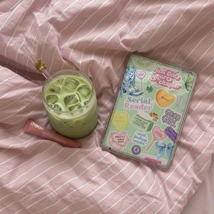 May include: A refreshing green beverage in a clear glass with ice cubes, a pink lip product, and a phone case with stickers on a pink striped bedsheet. The phone case has the text "Just One More Chapter".