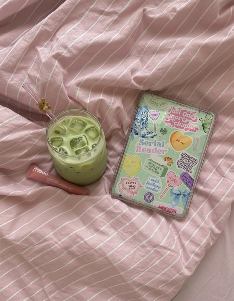 May include: A refreshing green beverage in a clear glass with ice cubes, a pink lip product, and a phone case with stickers on a pink striped bedsheet. The phone case has the text "Just One More Chapter".