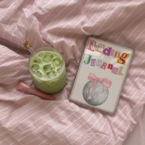 May include: A refreshing matcha latte with ice cubes in a clear glass, a lip product, and a "Reading Journal" with a disco ball illustration and pink bow, all on pink striped bedding.