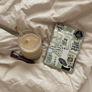 May include: A clear glass of coffee with a straw and a small bear decoration, a tube of rhode lip treatment, and a phone case with stickers on a bed. The phone case has the text "In my Kindle Era" and "Just one more page".