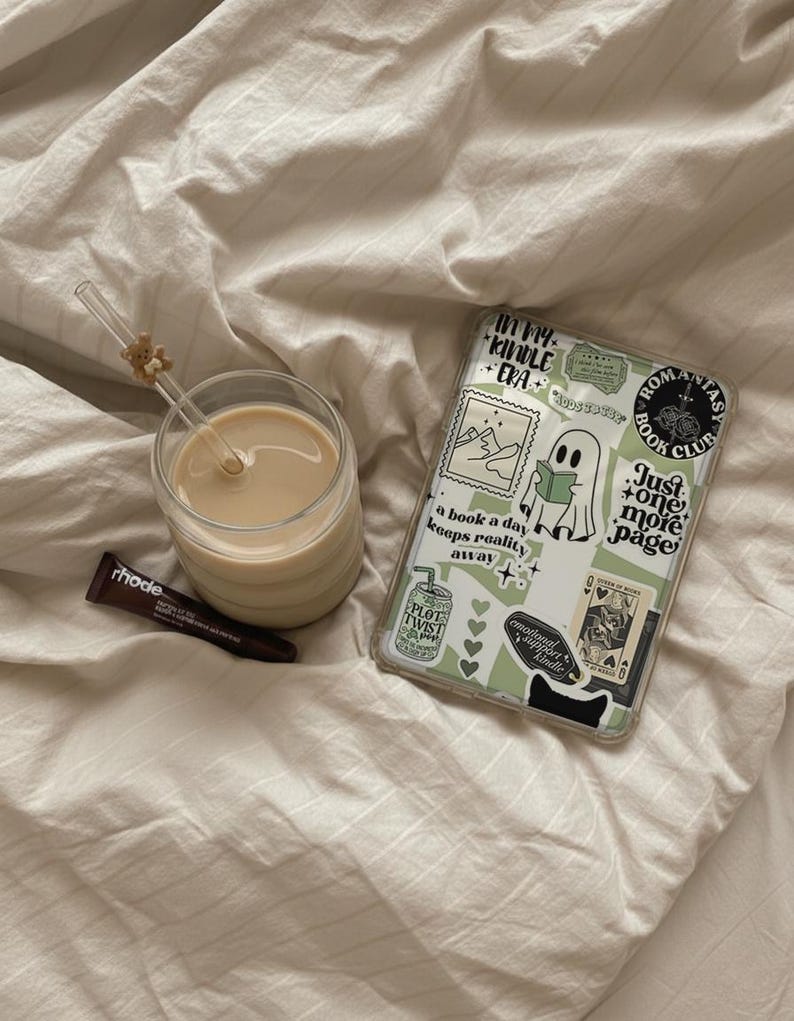 May include: A clear glass of coffee with a straw and a small bear decoration, a tube of rhode lip treatment, and a phone case with stickers on a bed. The phone case has the text "In my Kindle Era" and "Just one more page".