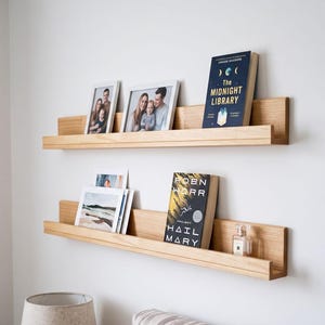 Puede incluir: Dos estantes flotantes de madera de tono claro montados en una pared blanca. El estante superior sostiene dos fotografías enmarcadas y un libro titulado The Midnight Library. El estante inferior muestra varias fotografías y un libro titulado Project Hail Mary.