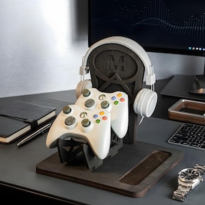 May include: A dark wood gaming organizer stand featuring a central vertical support with an engraved letter M, designed to hold two white game controllers in the front and a pair of white over-ear headphones on the side arms of the structure.