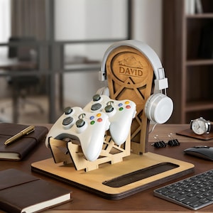 Op de afbeelding: Een lichtbruine houten gaming-organizerstandaard met een verticale steun voorzien van de gegraveerde naam DAVID en een bergafbeelding. De standaard houdt twee witte gamecontrollers en een witte hoofdtelefoon vast op een platte basis.