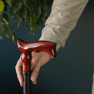 Op de afbeelding: Een houten wandelstok met een donkerrode, gepolijste handgreep. De handgreep heeft een uniek, gebogen ontwerp.