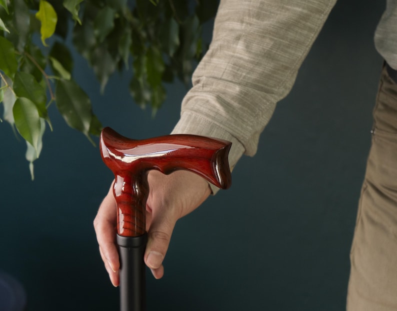 Op de afbeelding: Een houten wandelstok met een donkerrode, gepolijste handgreep. De handgreep heeft een uniek, gebogen ontwerp.