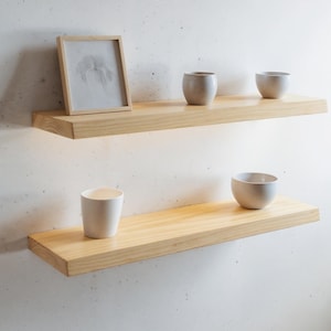 May include: Two light-coloured wooden floating shelves mounted on a white wall. The top shelf holds a framed picture and two small, grey ceramic cups. The bottom shelf displays a white ceramic cup and a grey bowl. The shelves are made of light-coloured wood.
