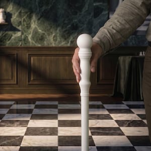 May include: A white walking cane with a rounded top and a decorative column design. The cane is held by a person in a room with a checkered floor and wood paneling. The cane's height is approximately 36 inches.