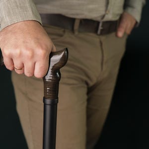 Op de afbeelding: Een persoon die een donkerbruine houten wandelstok vasthoudt met een zwarte metalen schacht.
