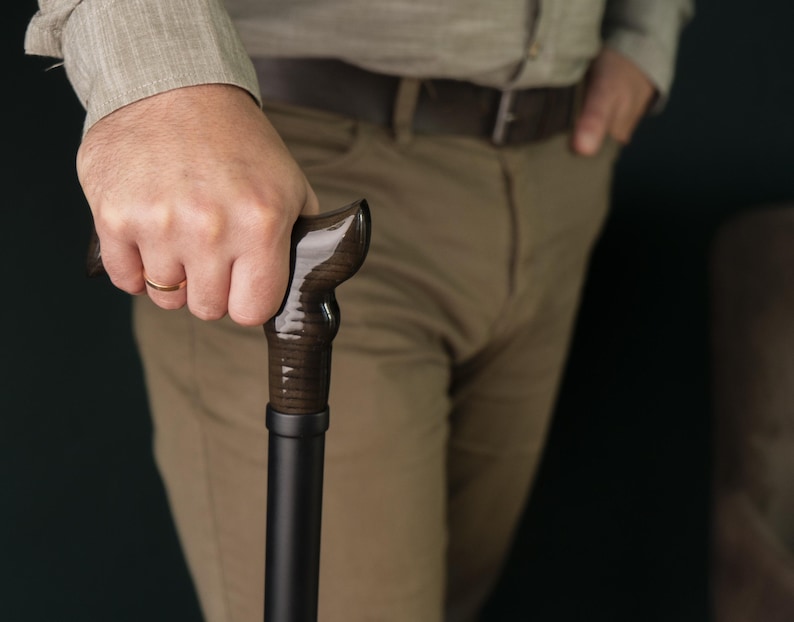 Op de afbeelding: Een persoon die een donkerbruine houten wandelstok vasthoudt met een zwarte metalen schacht.
