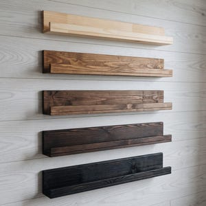 Puede incluir: Cinco estantes de pared horizontales de madera dispuestos en una pila vertical. Cada estante cuenta con un panel trasero plano y un borde frontal estrecho. Los acabados de madera van desde un tono natural claro hasta tintes oscuros.