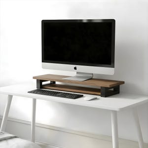 May include: A wooden monitor stand with a black base supports a silver computer monitor. A black keyboard and white mouse sit on a white desk. The stand has a lower shelf for storage.