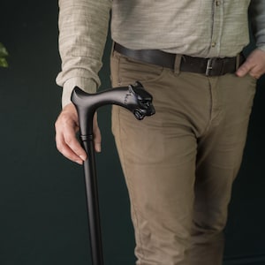 Puede incluir: Bastón negro con un mango decorativo con forma de cabeza de pantera. La persona lleva una camisa verde claro y pantalones color caqui. El fondo es una pared verde oscuro.