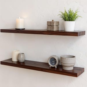 May include: Two horizontal dark brown wooden floating shelves mounted on a wall. The top shelf holds a white pillar candle, a stacked grey stone block, and a green plant in a white pot. The bottom shelf holds a white candle, a grey cup, a clock, and a bowl.