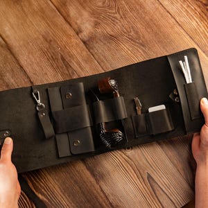 May include: Black leather pipe roll with multiple compartments. The roll contains a pipe, cleaning tools, and a lighter. The roll is open, displaying the contents, and is held by two hands on a wooden surface.