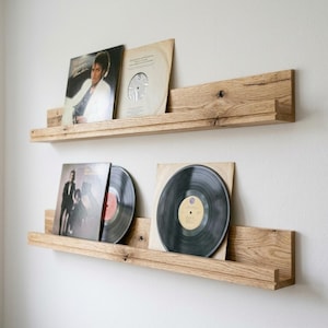 Könnte beinhalten: Zwei horizontale, an der Wand montierte Schweberegale aus hellem Eichenholz. Die Regale verfügen über eine erhöhte Vorderkante zum Abstellen von Gegenständen. Auf den Regalen stehen vier Schallplatten, zwei Plattencover und zwei schwarze Platten.