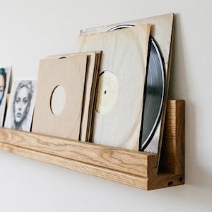 May include: A light brown wooden wall-mounted shelf holding several vinyl records in plain tan paper sleeves and a black and white portrait print. The shelf features a horizontal ledge and a vertical back panel to support the items upright.