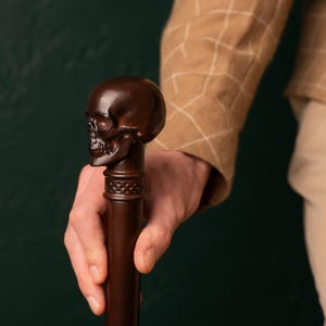 May include: A dark brown wooden walking stick with a carved skull head.