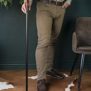 Op de afbeelding: Een persoon die een houten wandelstok vasthoudt met een donkerbruine handgreep en een zwarte schacht. De stok is ongeveer 90 cm lang. De persoon draagt ​​een bruine broek en bruine schoenen.