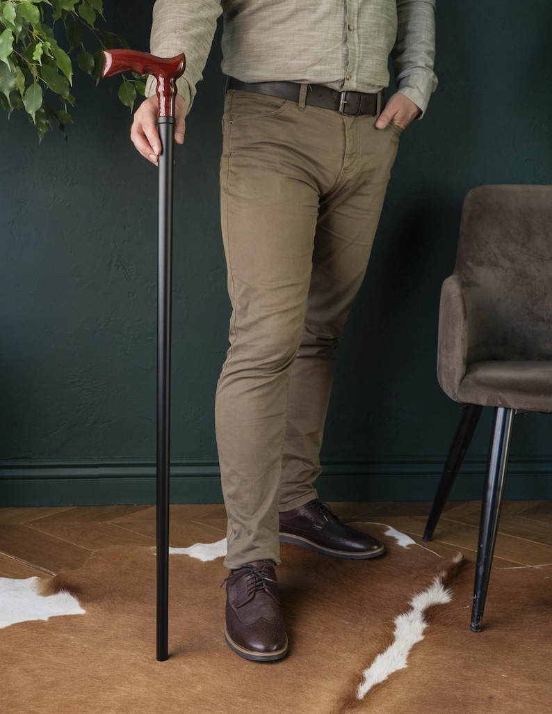Op de afbeelding: Een persoon die een houten wandelstok vasthoudt met een donkerbruine handgreep en een zwarte schacht. De stok is ongeveer 90 cm lang. De persoon draagt ​​een bruine broek en bruine schoenen.