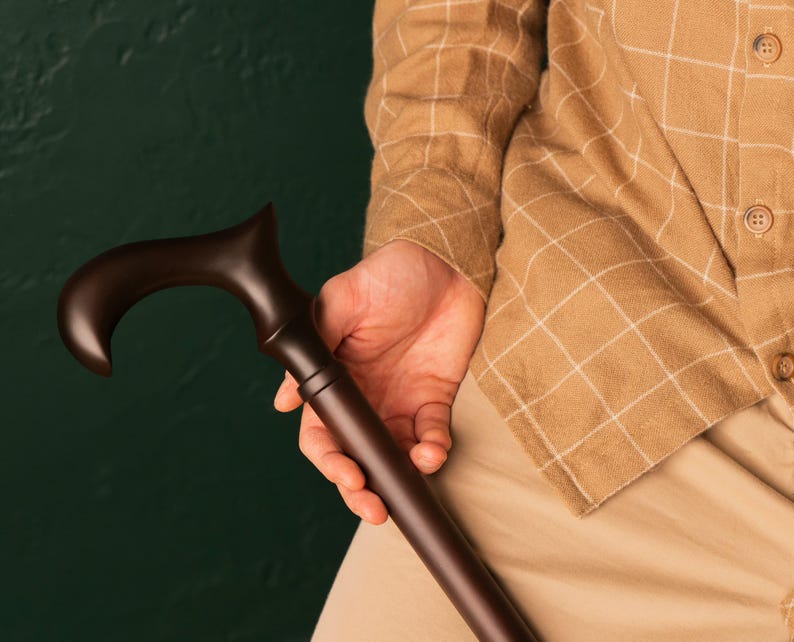 Puede incluir: Un bast&oacute;n de madera marr&oacute;n oscuro con un mango curvo. El bast&oacute;n lo sostiene una persona que lleva una camisa de cuadros marr&oacute;n claro.