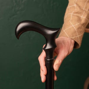 Könnte beinhalten: Ein schwarzer Holzspazierstock mit gebogenem Griff. Der Griff wird von einer Person gehalten, die eine braune Jacke trägt. Der Stock hat eine glatte, polierte Oberfläche und ein klassisches Design. Der Stock ist etwa 91 cm lang.