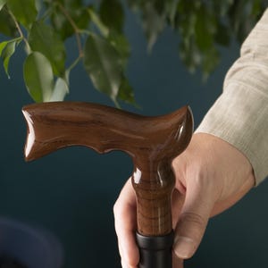 Op de afbeelding: Een bruine houten wandelstok met een gebogen handvat. Het handvat is glad en gepolijst, en de stok is gemaakt van donker hout.