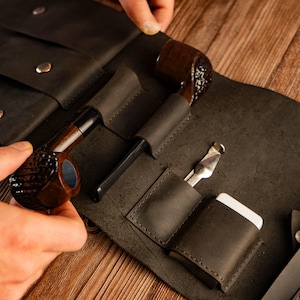 May include: A dark gray leather pipe roll with two pipes, a tamper, and a cleaning tool. The pipes are brown with black stems. The roll has pockets and snap closures. The background is a wooden surface.