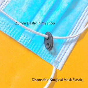 May include: Close-up of a blue disposable surgical mask with white elastic ear loops. The elastic is threaded through a black plastic adjuster. The text "2.5mm Elastic in my shop" is visible.