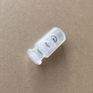 Peut inclure: Un bouton de r&eacute;glage en plastique transparent avec un ressort &agrave; l'int&eacute;rieur. Le bouton a deux trous de chaque c&ocirc;t&eacute; pour un cordon ou une ficelle.