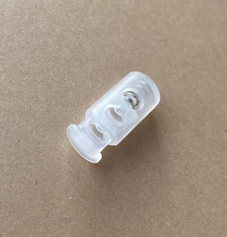 Peut inclure: Un bouton de r&eacute;glage en plastique transparent avec un ressort &agrave; l'int&eacute;rieur. Le bouton a deux trous de chaque c&ocirc;t&eacute; pour un cordon ou une ficelle.