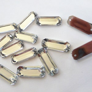 May include: A group of silver-toned rectangular glass rhinestones. The rhinestones have a faceted design and are scattered on a white surface.