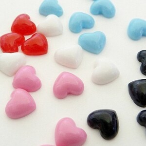 May include: A collection of small, smooth, heart-shaped cabochons in various solid colors including red, white, light blue, pink, and black. Each heart features a glossy, rounded surface and a flat back, arranged in a scattered pattern.
