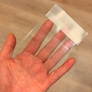 May include: A clear, rectangular cellophane bag with a white adhesive strip across the top. The bag is held in a person's hand, with the fingers visible through the transparent material. The background is a light brown wooden surface.
