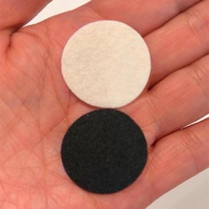 May include: Two circular felt pads, one cream-colored and one black, are displayed in a hand. The pads are of similar size and shape, with a slightly textured surface. The background is a light skin tone.