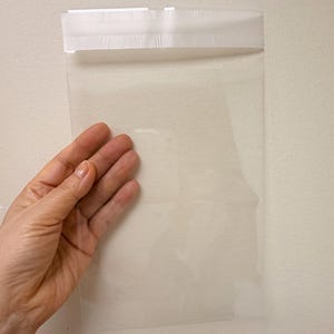Op de afbeelding: Een doorzichtige, rechthoekige plastic zak met een witte plakstrip aan de bovenrand om de zak af te sluiten. De zak is transparant, waardoor de inhoud volledig zichtbaar is, en is ontworpen voor het veilig verpakken van diverse items.