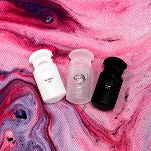 Könnte beinhalten: Drei Kunststoff-Kordelstopper in Weiß, Klar und Schwarz. Die Kordelstopper liegen auf einer rosa-weißen Marmorfläche.