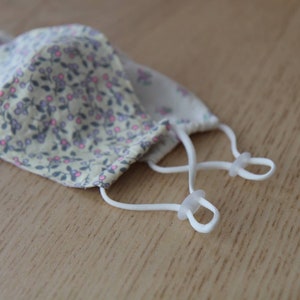 May include: A white face mask with a floral pattern and white elastic straps with plastic adjusters.