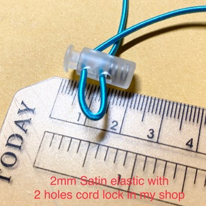 May include: Close-up of a clear plastic cord lock with two holes, measuring 2 cm in length, holding a turquoise elastic cord. The cord lock is shown against a ruler.