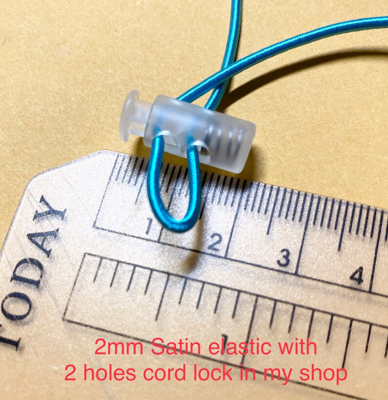 May include: Close-up of a clear plastic cord lock with two holes, measuring 2 cm in length, holding a turquoise elastic cord. The cord lock is shown against a ruler.