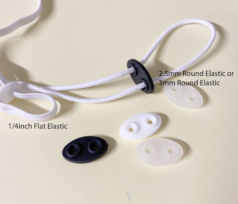 May include: White elastic cord with two white plastic slider toggles and one black plastic slider toggle. The elastic cord is labeled "1/4inch Flat Elastic". One of the white toggles is labeled "2.5mm Round Elastic or 3mm Round Elastic".