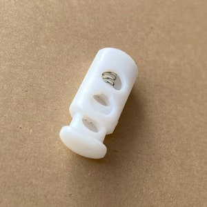 Peut inclure: Toggle en plastique blanc avec un ressort &agrave; l'int&eacute;rieur et trois trous. Le toggle est utilis&eacute; pour ajuster la longueur des cordons ou des sangles.