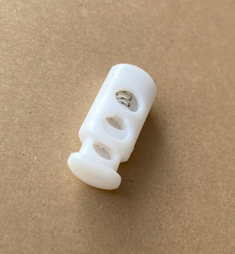 Peut inclure: Toggle en plastique blanc avec un ressort &agrave; l'int&eacute;rieur et trois trous. Le toggle est utilis&eacute; pour ajuster la longueur des cordons ou des sangles.