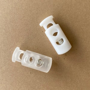 Peut inclure: Deux bloqueurs de cordon en plastique blanc avec un ressort m&eacute;tallique &agrave; l'int&eacute;rieur. Les bloqueurs de cordon sont utilis&eacute;s pour ajuster la longueur des cordons ou des sangles.