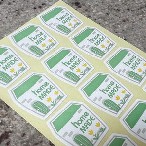 May include: A sheet of stickers with a house design. Each sticker features a white house with a green roof and door, the words "home MADE" in green, and yellow flower accents. The stickers are on a light yellow background.